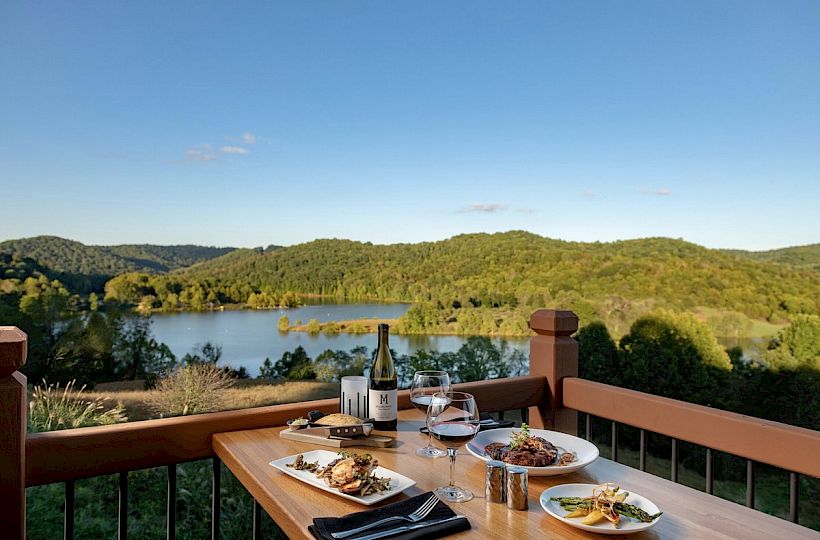 A dining table set with food and wine overlooks a serene lake surrounded by lush green hills under a clear blue sky.
