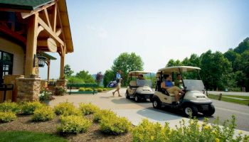 The image shows two people in golf carts near a clubhouse, with another person carrying a golf bag towards the building. There are flowers in the foreground.