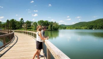 Two people stand on a wooden boardwalk overlooking a calm lake, surrounded by lush greenery and hills under a clear blue sky.