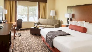 The image shows a hotel room with a large bed, a desk with a chair, a TV, a sofa, lamps, and a window letting in light, ending the sentence.