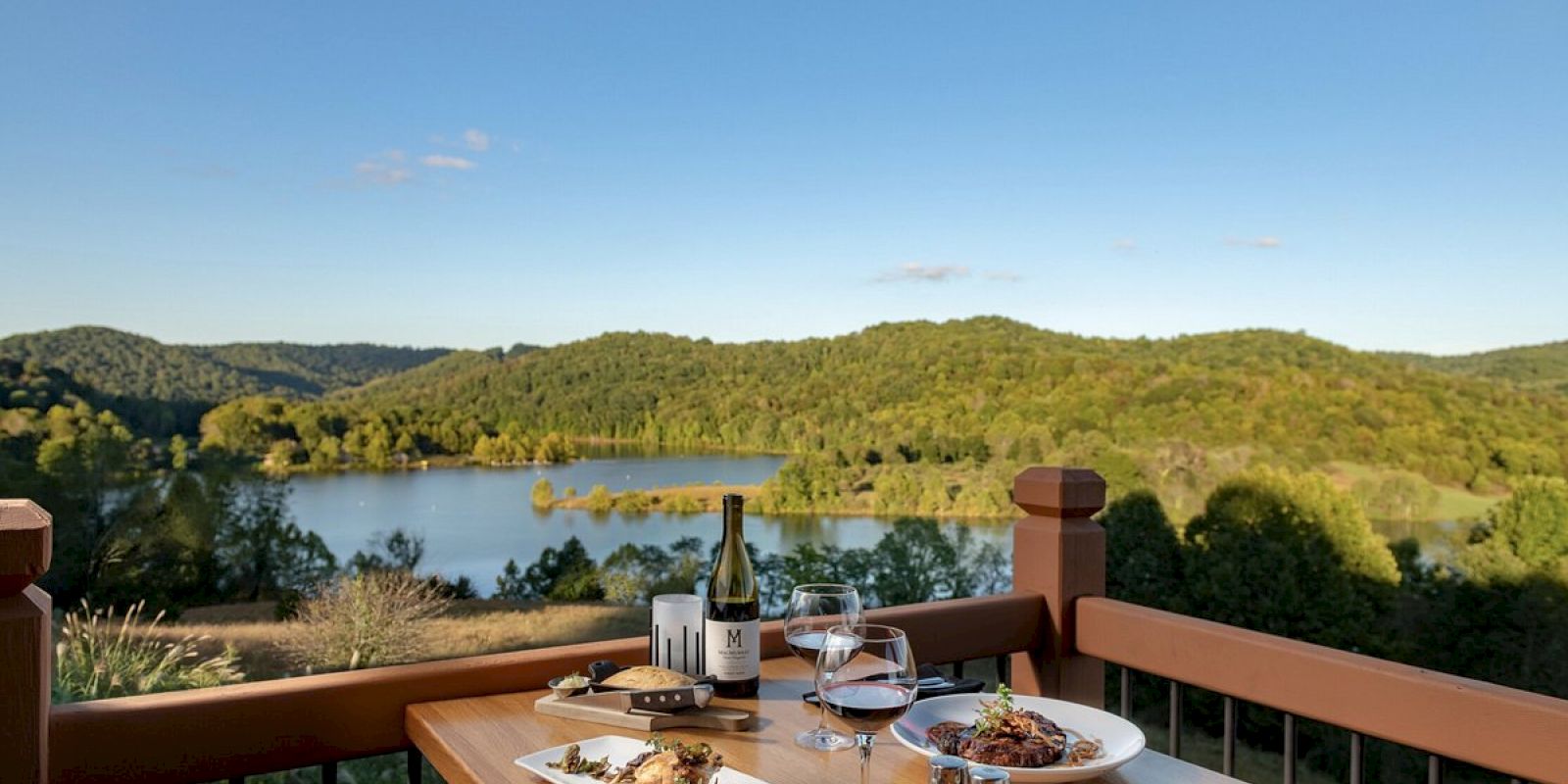 A table set with food and wine overlooks a scenic lake surrounded by lush green hills under a clear blue sky from a wooden deck railing.