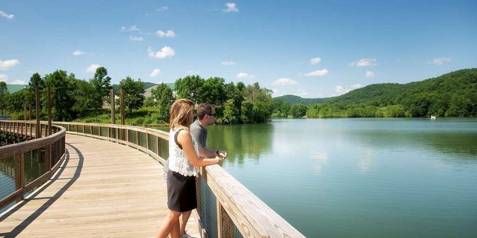 A man and a woman stand on a wooden walkway by a serene lake, surrounded by green hills and trees under a clear blue sky.