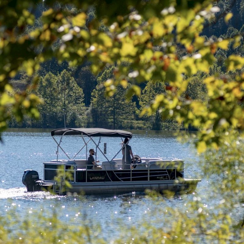 A pontoon boat with two people is cruising on a lake, surrounded by lush greenery with trees framing the scene.