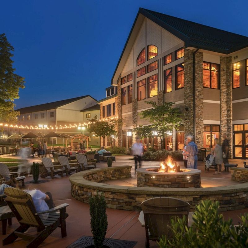 An evening outdoor scene with a large building, lit string lights, a fire pit surrounded by people on lounge chairs, and a lush garden environment.