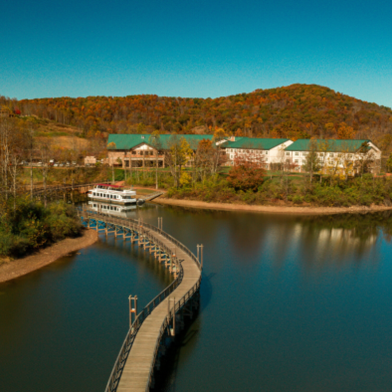 A peaceful lakeside scene with a curved bridge, docked boat, and buildings surrounded by autumn foliage, under a clear blue sky.