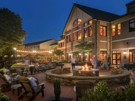 An outdoor evening scene of a cozy lodge with people sitting by a circular fire pit, surrounded by string lights and Adirondack chairs.
