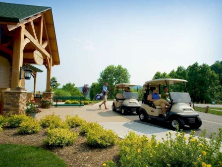 The image shows people with golf carts near a building with wooden beams, surrounded by greenery and yellow flowers on a sunny day.