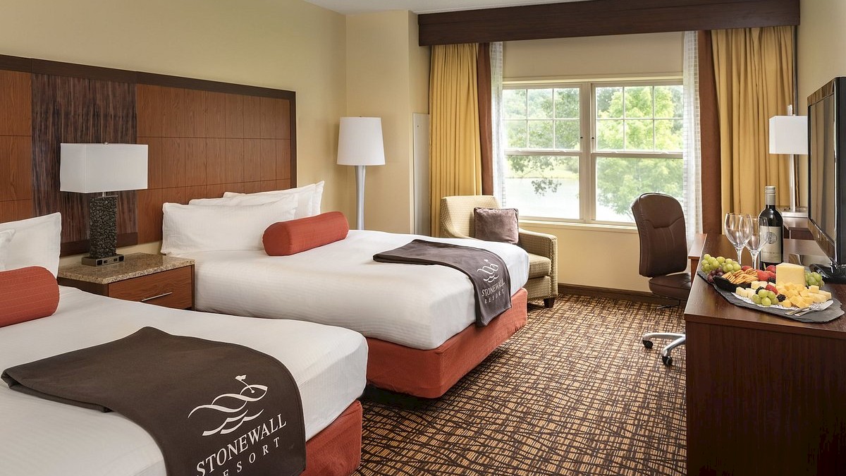 The image shows a well-appointed hotel room with two beds, a large window, a lamp, an armchair, a flat-screen TV, and a plate of fruit and wine.