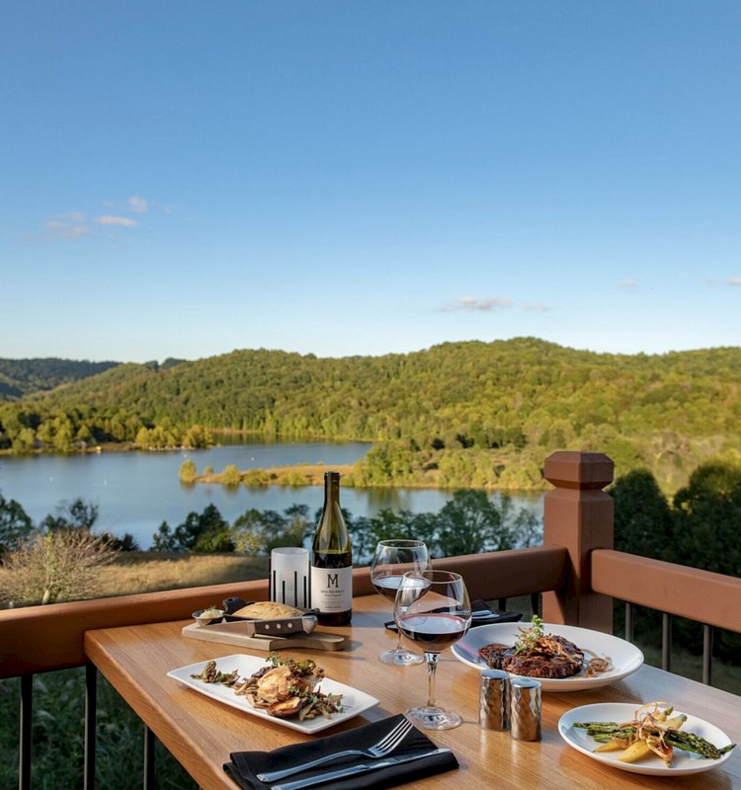A table with plates of food, wine, and glasses set on a balcony overlooking a serene lake and lush green hills on a clear day.