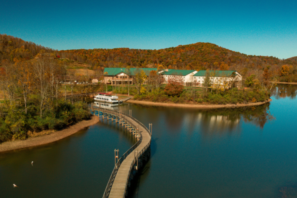 The image shows a scenic lakeside view with a boardwalk leading to buildings with green roofs, surrounded by trees and hills under a clear blue sky.