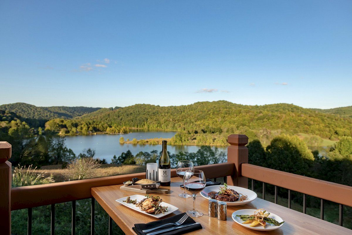A dining table with food, wine, and water is set on a balcony overlooking a serene lake with surrounding green hills and a clear blue sky.