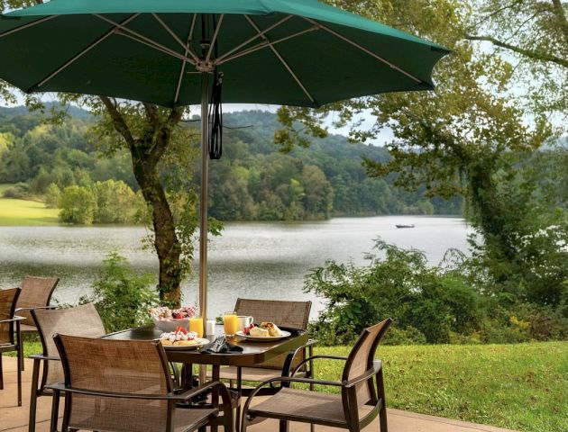 Outdoor dining table with chairs, green umbrella, and breakfast food, set near a scenic lake with trees and hills in the background, boat on water.