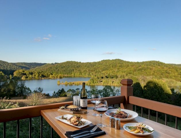 A table set with food and wine overlooks a beautiful landscape with a lake and green hills under a clear blue sky, providing a serene dining experience.