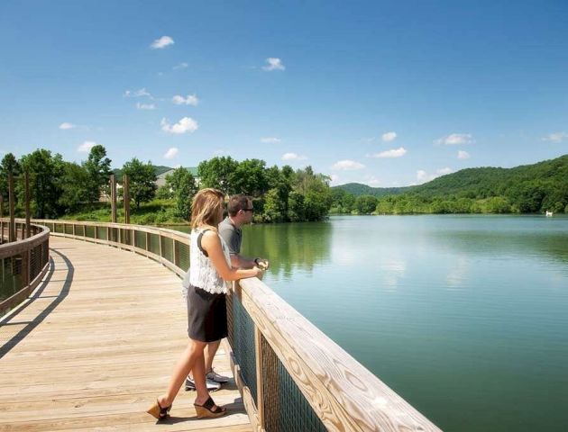A couple stands on a wooden walkway overlooking a serene lake surrounded by lush green hills under a clear, blue sky with a few scattered clouds.