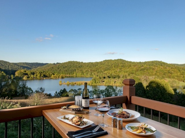 A table set for a meal on a balcony overlooks a scenic lake and forested hills, with dishes, wine, and glasses arranged on the table.