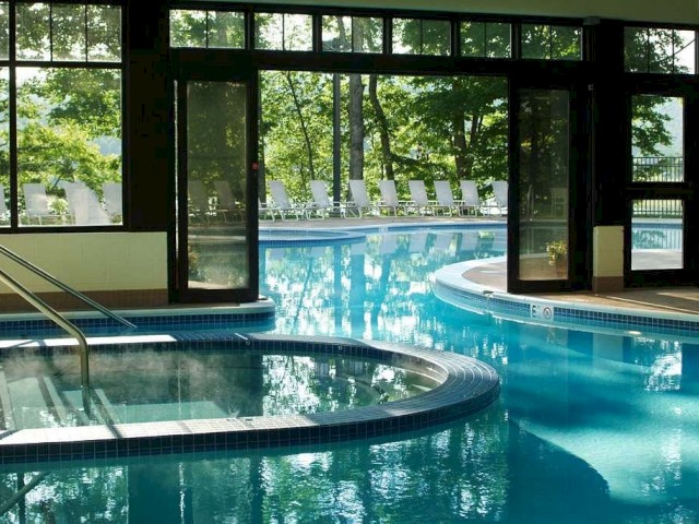 The image shows an indoor swimming pool area with large glass windows and doors, connecting to an outdoor pool surrounded by lush greenery and lounge chairs.