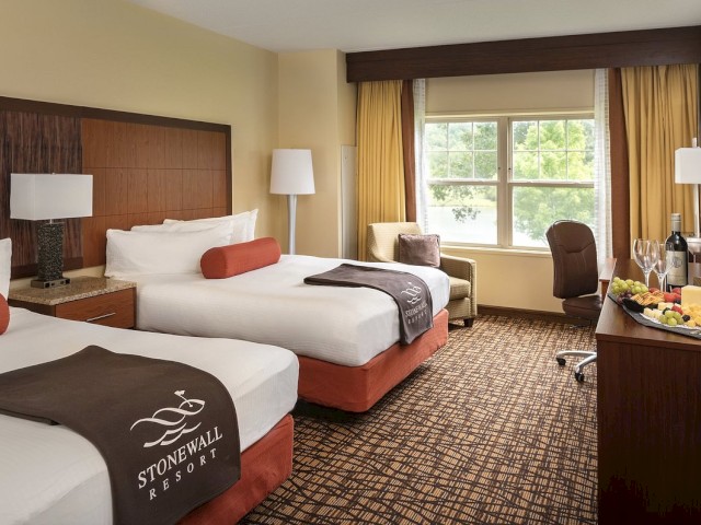 A hotel room with two beds, a chair by the window, a dresser with wine and fruit, and a view outside. The blankets say 