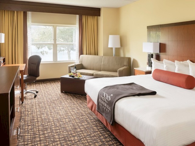 A hotel room with a large bed, sofa, desk, chair, TV, and two lamps. The room has a window with curtains drawn, a simple yet elegant decor.