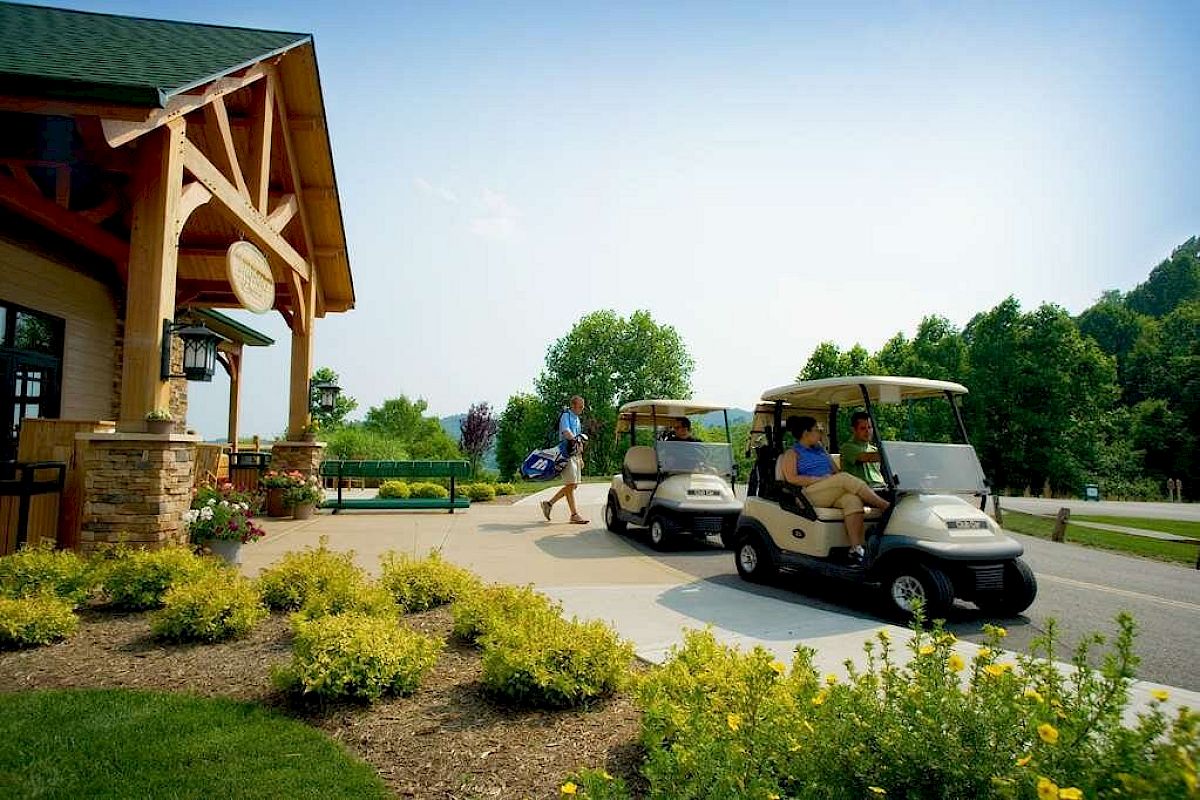 A person walks with a golf bag near a building, while two people sit in golf carts on a paved area surrounded by greenery and landscaping.