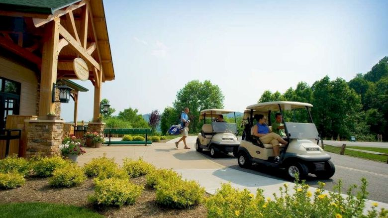 A person walks with a golf bag near a building, while two people sit in golf carts on a paved area surrounded by greenery and landscaping.