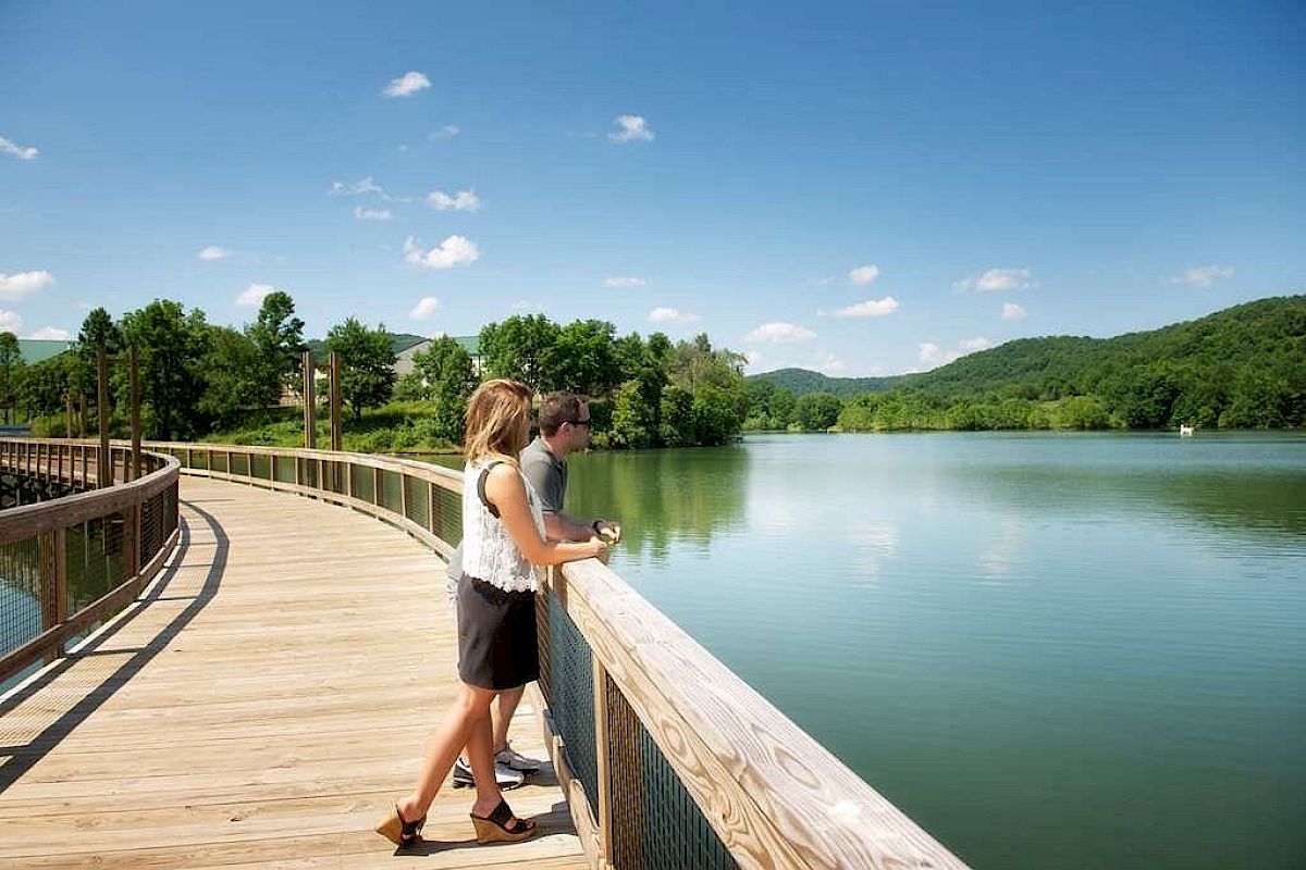 A man and woman stand on a wooden boardwalk overlooking a calm lake, surrounded by lush green hills and blue skies with scattered clouds.