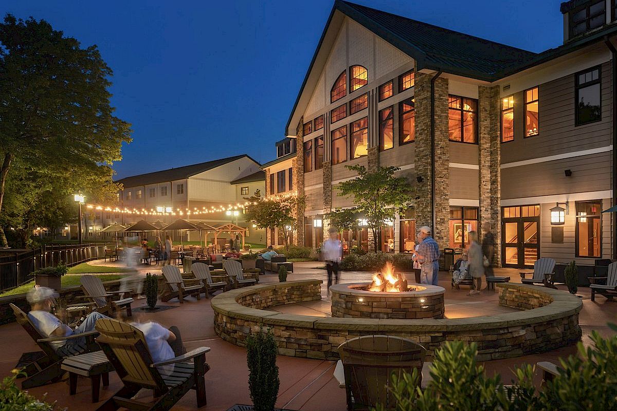 An inviting outdoor lounge area at dusk with a fire pit, casual seating, string lights, and people enjoying the atmosphere near a large building.
