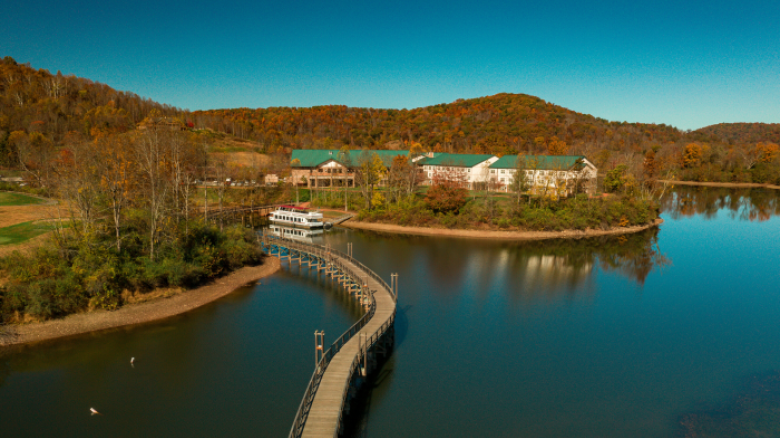 The image shows a scenic lakeside area with buildings, a boat docked at a curved pier, and surrounding hills with autumn foliage under a clear blue sky.