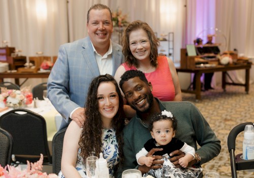 A diverse group of five people, including a baby, smiling and posing at a festive dinner event with elegant decor and flowers.