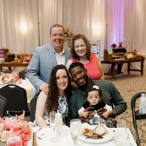A diverse group of five people, including a baby, smiling and posing at a festive dinner event with elegant decor and flowers.