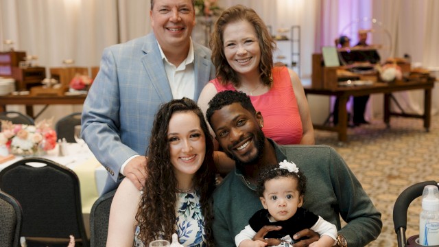 A diverse group of five people, including a baby, smiling and posing at a festive dinner event with elegant decor and flowers.