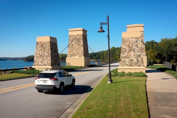 A white SUV drives past three stone columns on a bridge over a lake on a sunny day.